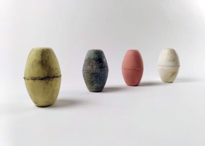 Photo de 4 flotteurs de filet de pêche alignés. Le flotteurs de gauche le plus proche et la ligne s'éloignant. Les 3 premiers flotteurs sont des reproduction en céramique du dernier flotteur qui est l'original en plastique blanc. De gauche à droite les flotteurs sont jaune, vert puis orange clair. Ils portent tous les traces comme s'ils avaient été beaucoup utilisés et usés par le temps.