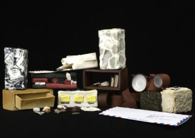 Briques en céramique inspirées de divers objets (tampons de passeport, ceinture en cuir, vêtement pliés, médicaments) placés en arc de cercle. Quelques coquillages et petites cailloux trouvés sur la plage. Une enveloppe.