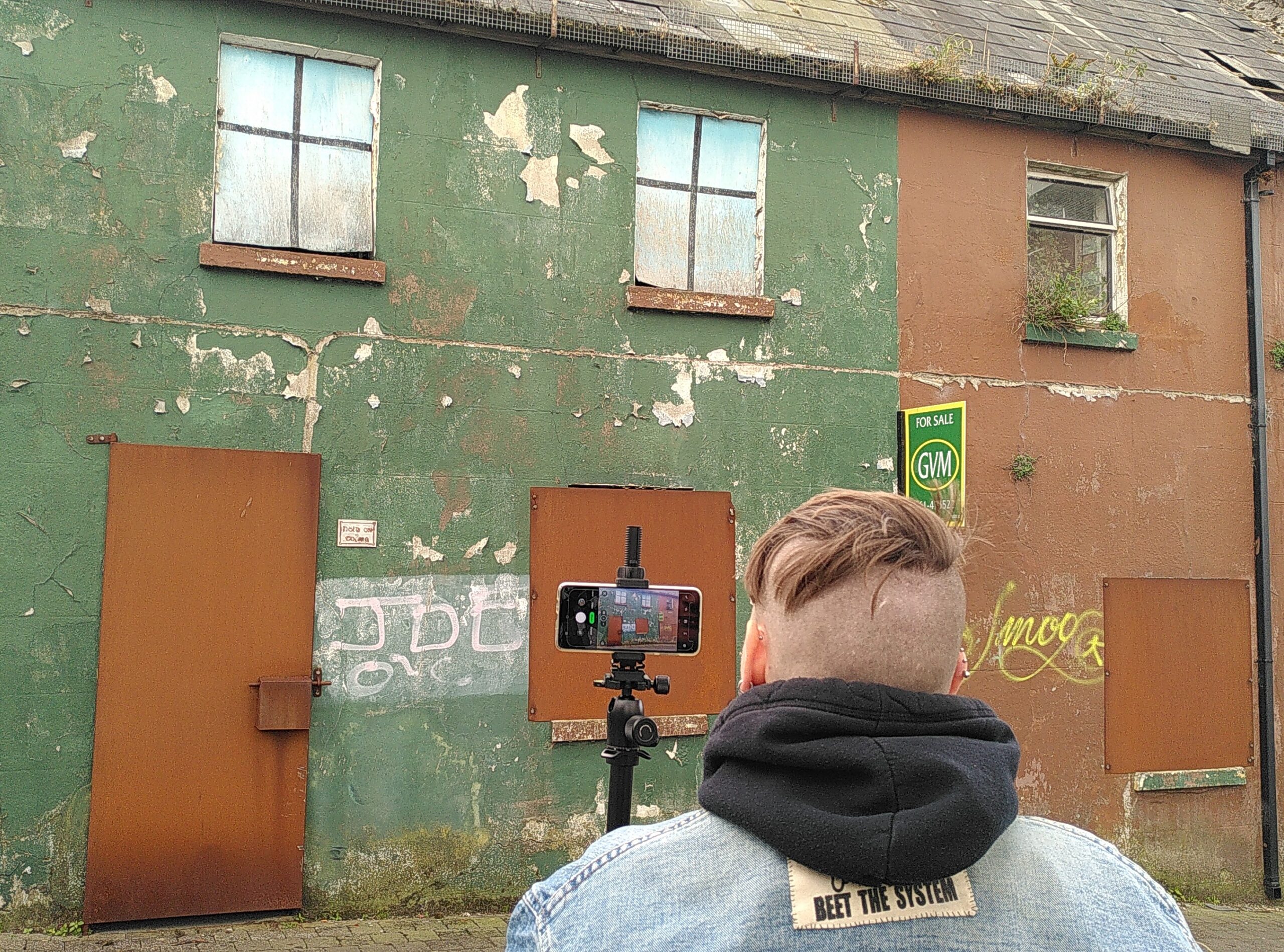 Charlie (Lauréline) Yris est de dos en train de prendre une photo d'une maison délabrée. Charlie porte une veste en jean et on voit un patch écrit "Beet the system" cousu dans le dos. Capuche d'un hoodie noir. Crâne rasé en undercut, cheveux courts. Les deux maisons sont fermées avec des plaque de métal. Peinture verte et rouge qui s'effrite, des plantes qui poussent sur les murs et fenêtres.