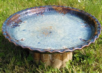 Une baignoire à oiseaux en céramique posée sur l'herbe dans un jardin. C'est un bassin en céramique avec un émail bleu plein de variations de teintes avec un pied pour le surélever. Il y a de l'eau dedans.