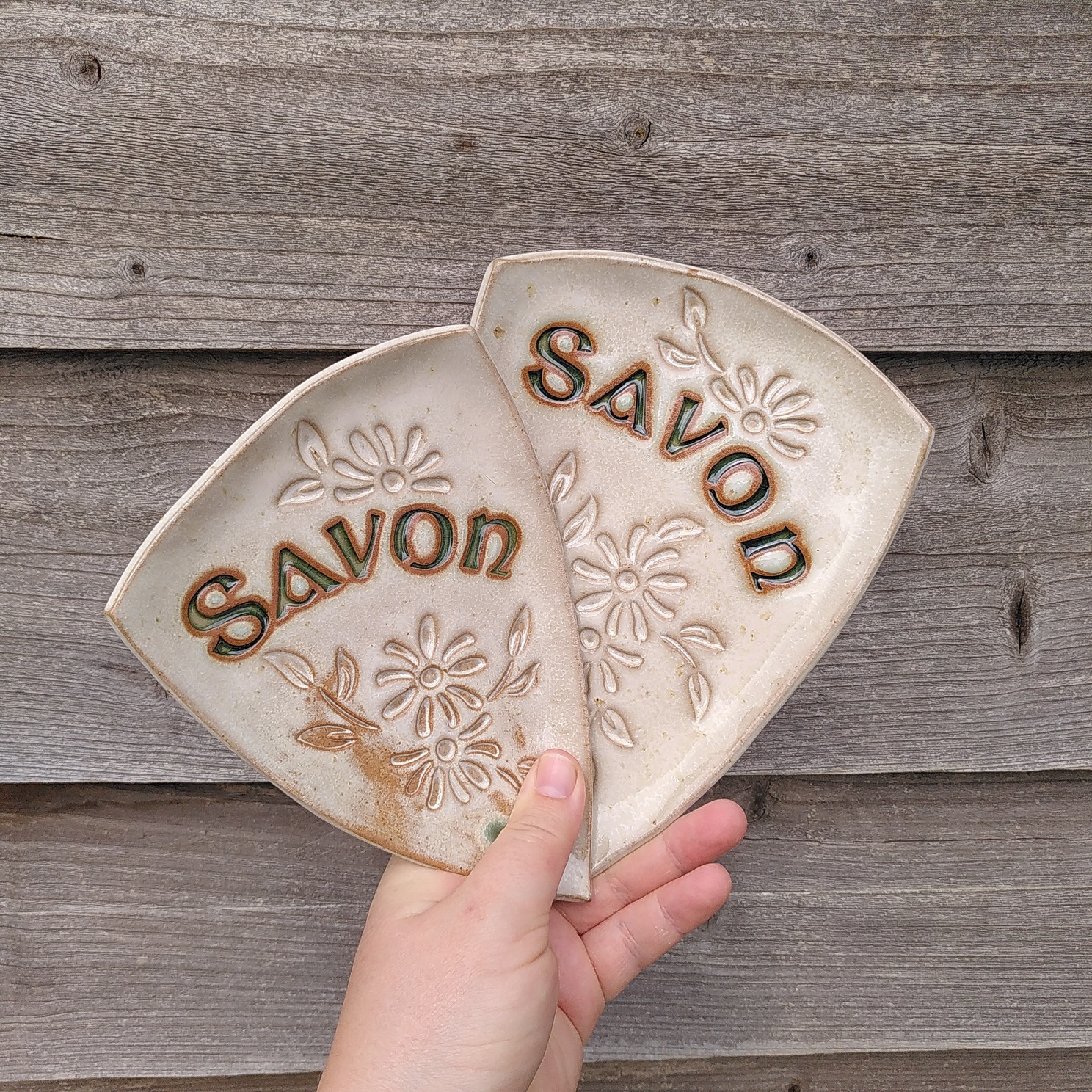 Deux porte-savons triangulaires en céramique émaillés en blanc cendre de bois. Il y a des fleurs et le mot savon estampés dessus.